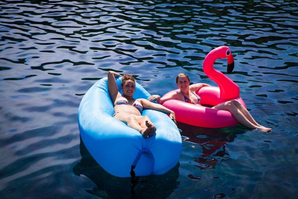 Zwei Menschen entspannen sich während ihres Segelurlaubs auf aufblasbaren Ringen, einer davon in Form eines blauen und einer in Form eines rosa Flamingos, und treiben auf dem klaren, blauen Wasser.