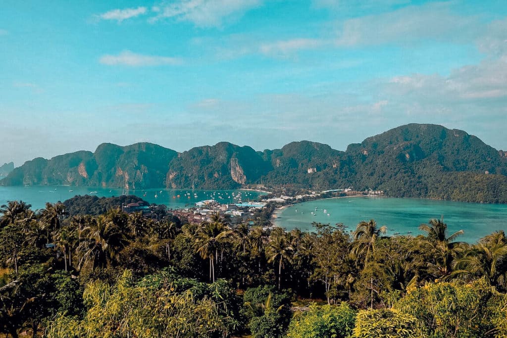 Eine malerische Aussicht auf ein Küstendorf, umgeben von üppigem, tropischem Wald und hoch aufragenden Kalksteinklippen mit Blick auf eine ruhige, türkisfarbene Bucht, perfekt für einen Segelurlaub.