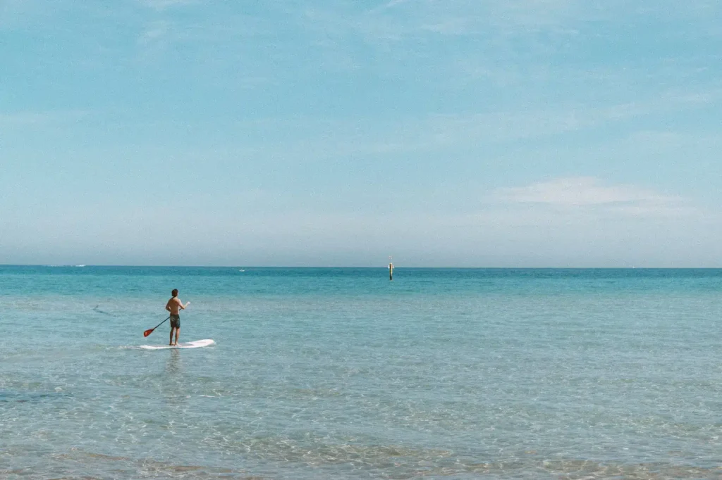 Eine Person fährt mit dem Paddleboard auf einem ruhigen, klaren Meer unter einem weiten blauen Himmel, mit einem dünn besiedelten Horizont und einer in der Ferne sichtbaren Segelyacht.