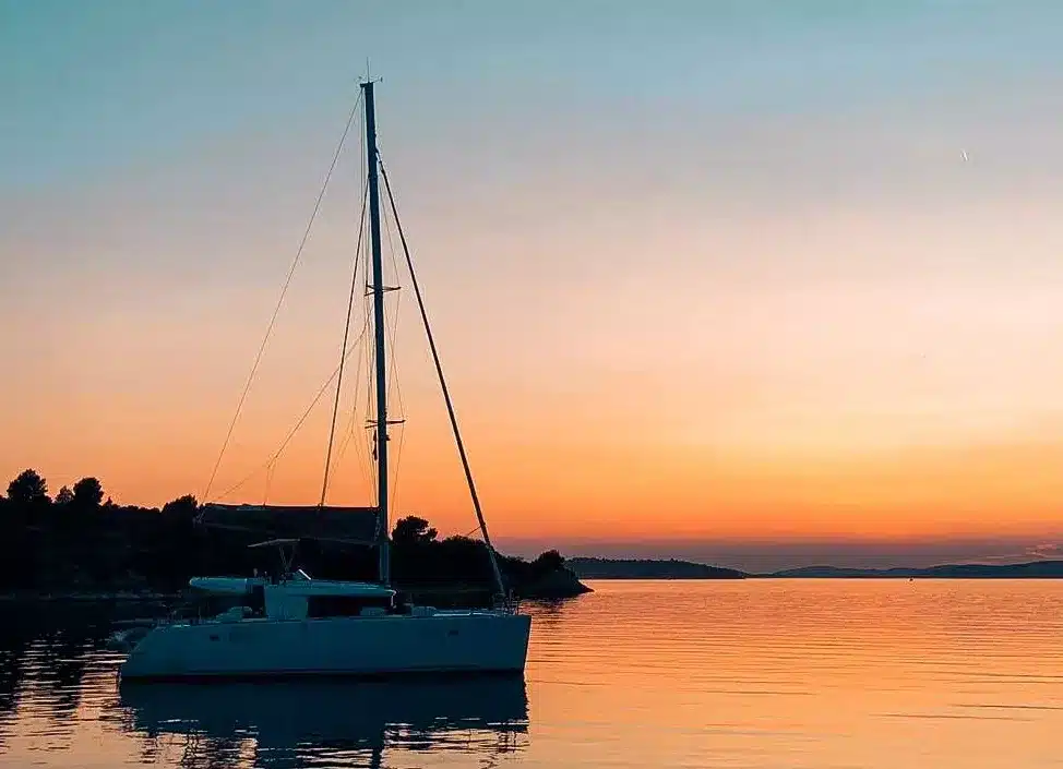 Eine Segelyacht auf ruhigen Gewässern während eines Sonnenuntergangs mit leuchtenden Orange- und Rosatönen am Himmel, die sich im Wasser nahe der Silhouette einer bewaldeten Küste spiegeln.