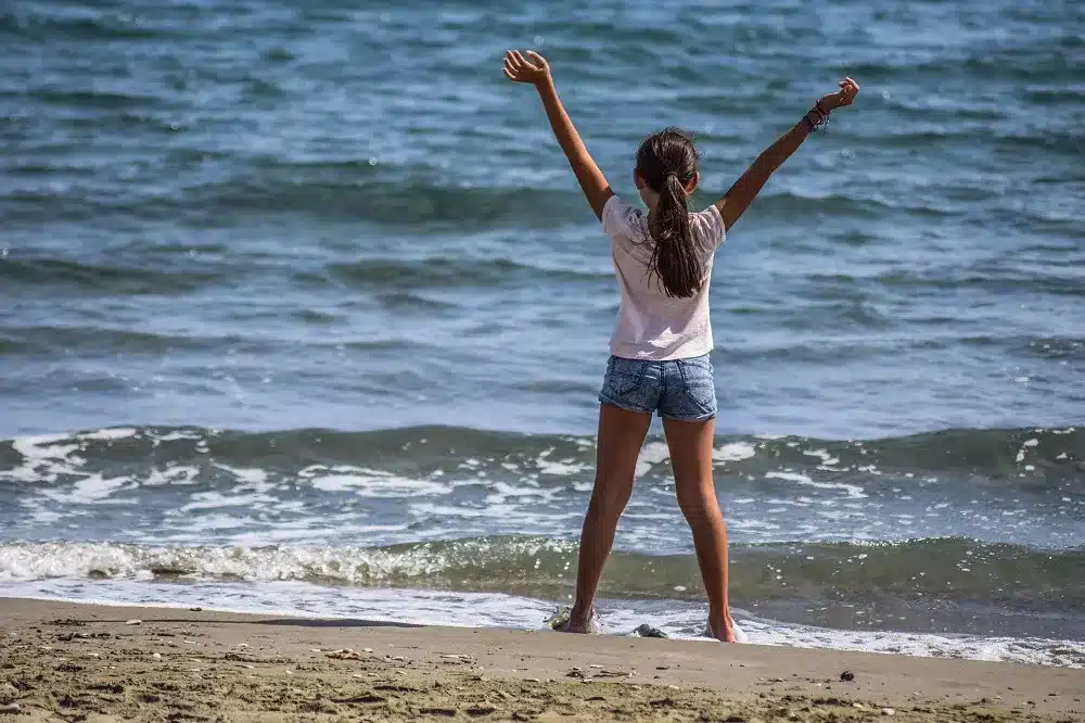 Ein junges Mädchen steht mit freudig erhobenen Armen an einem Sandstrand, blickt auf die Wellen des Ozeans und träumt von ihrer nächsten Segelreise.