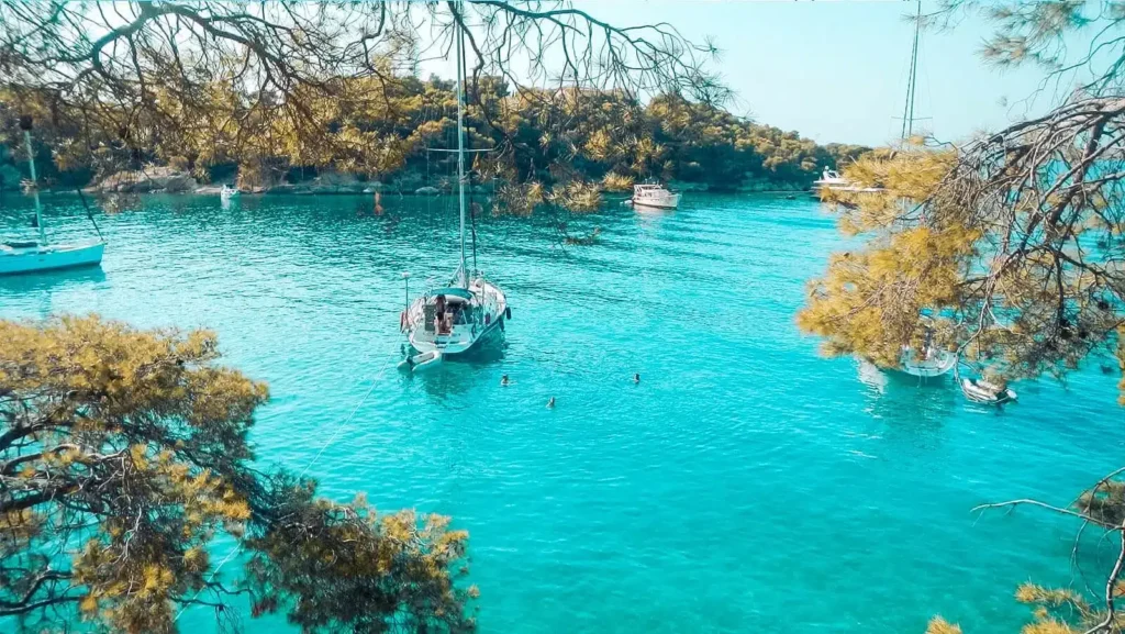 Eine ruhige Bucht mit türkisfarbenem Wasser, umgeben von üppigem Grün, wo zwei Segelboote vor Anker liegen und Menschen neben einem der Boote unter klarem Himmel schwimmen. Genießen Sie ein malerisches Segelt