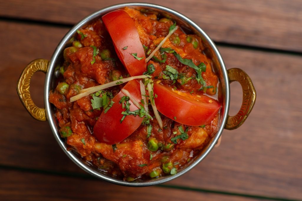 Eine Schüssel indisches Curry mit Erbsen, belegt mit frischen Tomatenscheiben und garniert mit Kräutern, serviert in einer dekorativen Metallschüssel auf einem Holztisch.