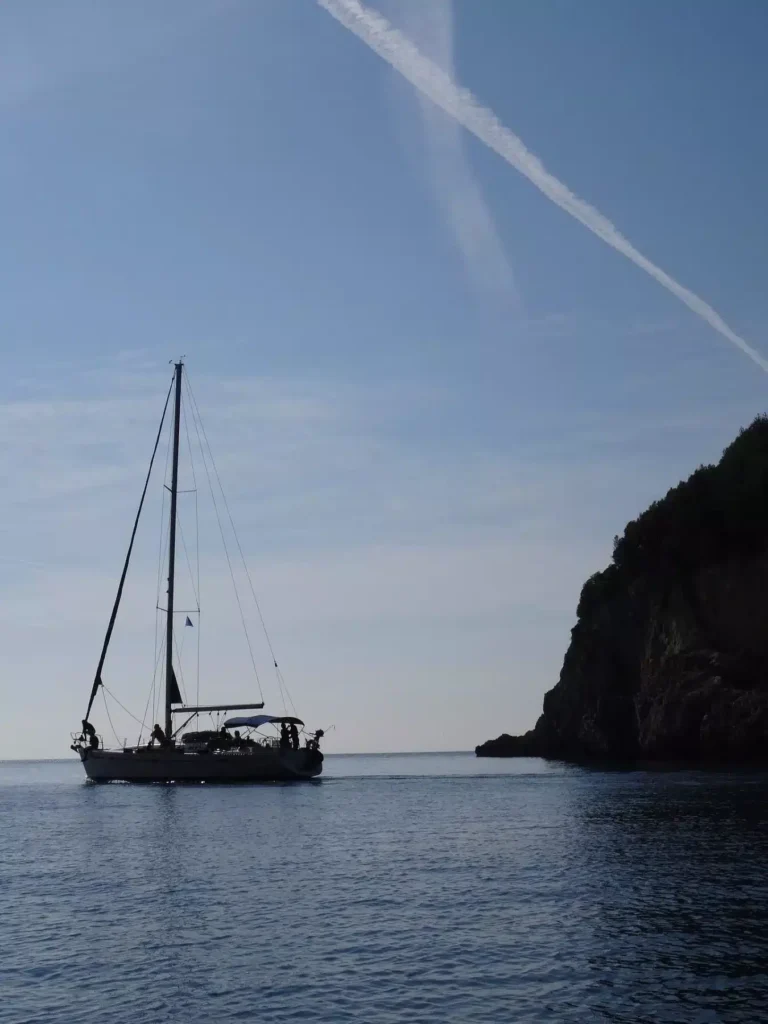 Eine Segelyacht liegt vor der Küste in der Nähe einer felsigen Küste unter einem klaren blauen Himmel. Am Himmel ist deutlich ein Kondensstreifen sichtbar.