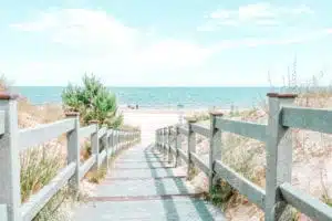 Ein Holzsteg mit Geländer führt durch Sanddünen zu einem ruhigen Strand unter einem strahlenden Himmel, ideal für den Aufbruch zu einem Segelabenteuer.