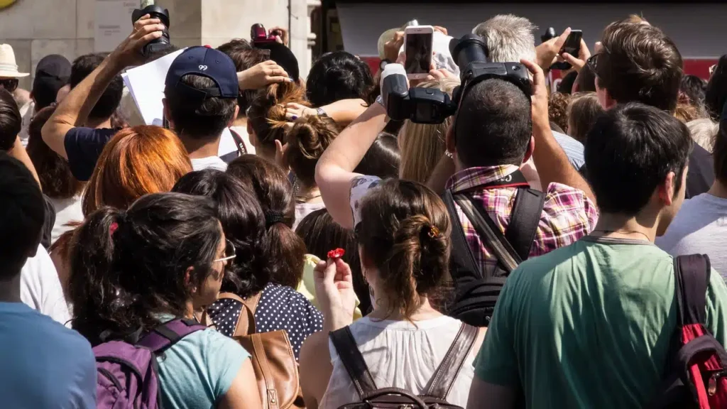 Eine Menschenmenge, von hinten und oben gesehen, viele halten Kameras und Smartphones hoch und fokussieren auf etwas außerhalb des Sichtfelds. Die Szene lässt auf eine geschäftige öffentliche Veranstaltung oder eine Touristenattraktion schließen, die von Übertourismus geplagt ist. Die Personen sind unterschiedlichen Alters und leger gekleidet.